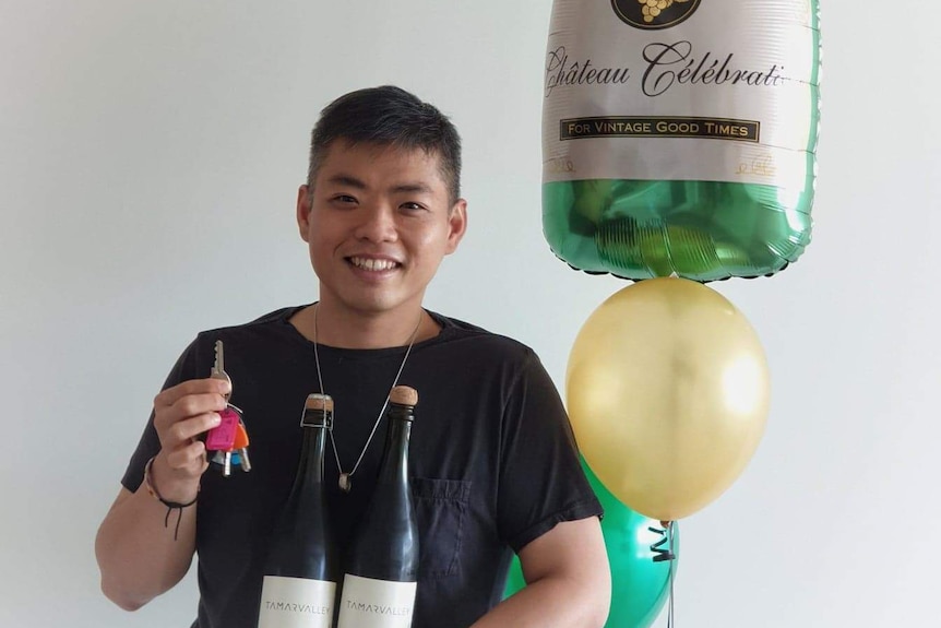A man smiles while holding a set of keys and two bottles of sparkling wine. There are balloons behind him