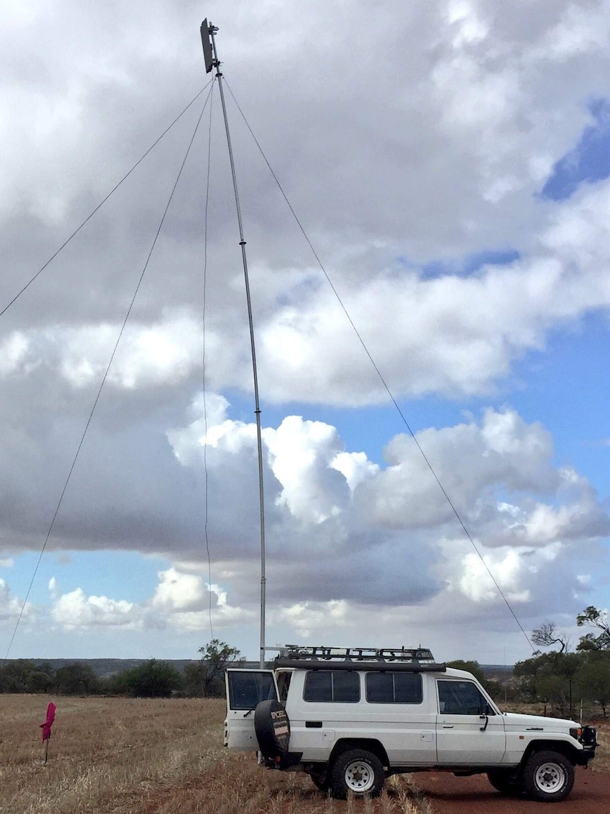 A white 4WD with a tall aerial-type tower attached to it with cables iis parked in the middle of a paddock on a cloudy day
