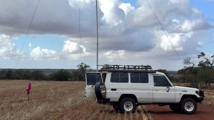 A white 4WD with a tall aerial-type tower attached to it with cables iis parked in the middle of a paddock on a cloudy day