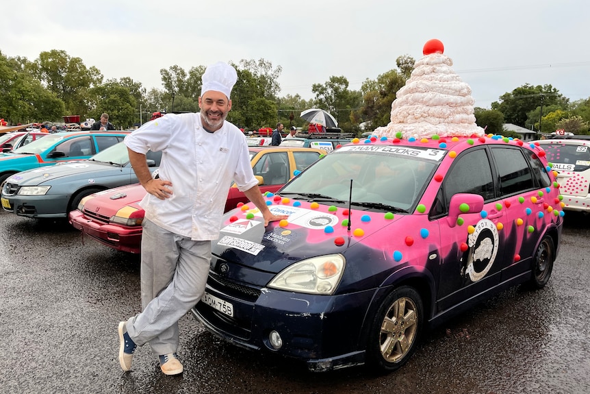 Man is a chefs outfit standing next to a pink car with a cake on the top