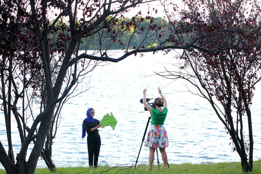 ABC camera operator filming Heywire winner holding green map of Australia while standing by a lake.