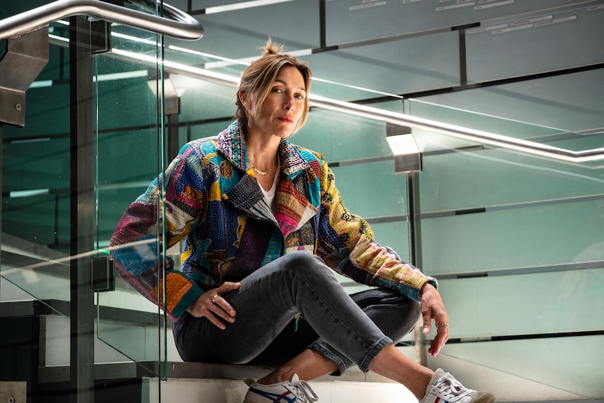 Karvan, in a multi-coloured and patterned jacket and jeans, sits atop some stairs in a modern glass-walled space.