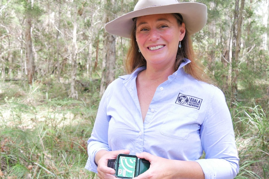 A woman wearing a hat and smiling at the camera, holding a small remote recording device.