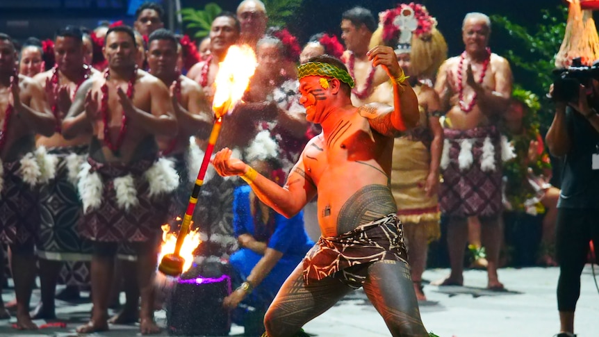 A man twirls a stick of fire while others watch on in the background.