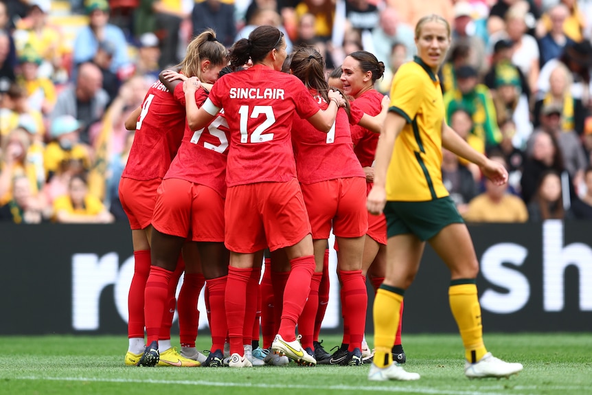 Canada players group together and hug as a Matildas play looks back over her shoulder
