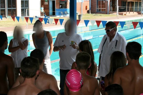 A group of coaches stand in front of young swimmers at a pool.