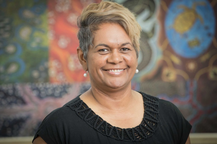 Headshot of an Indigenous woman with short blonde hair wearing a black top standing in front of Aboriginal artwork