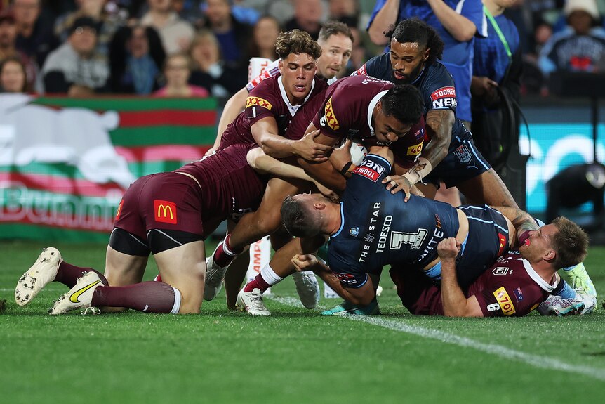 A NSW male State of Origin player holds the ball while he is held up over the goal line by Queensland.