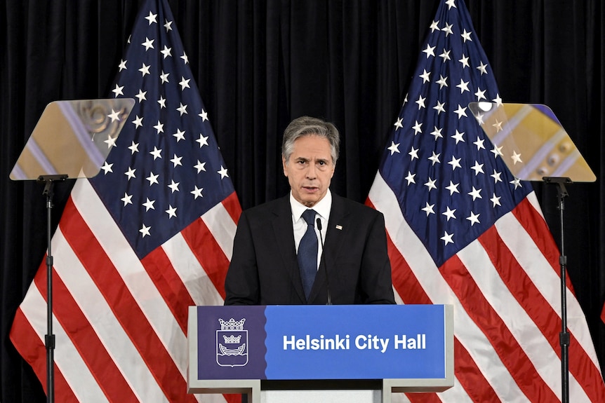 Antony Blinken stands between two US flags as he gives a speech from behind a lecturn with "Helsinki City Hall" on it.