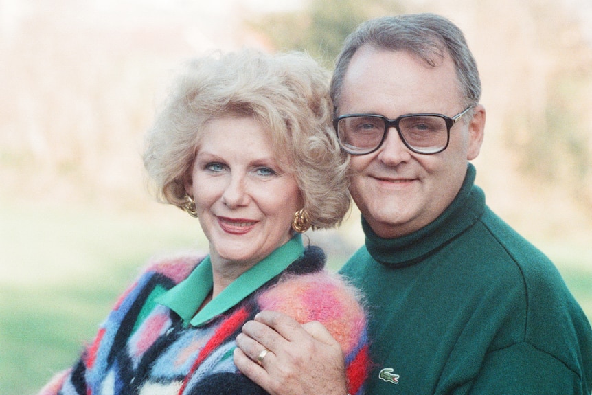 A man in a green jumper and glasses and woman in a colourful jumper standing together.