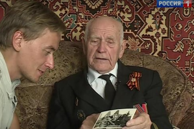 A man in a suit with medals adorning it shows a reporter to his left an old picture.