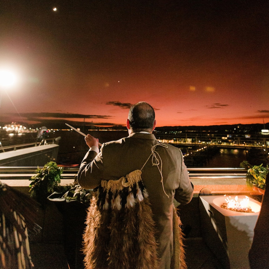 Man's back to camera, dressed in traditional Maori garment and suit overlooks city lights at first light. Dark and orange sky. 
