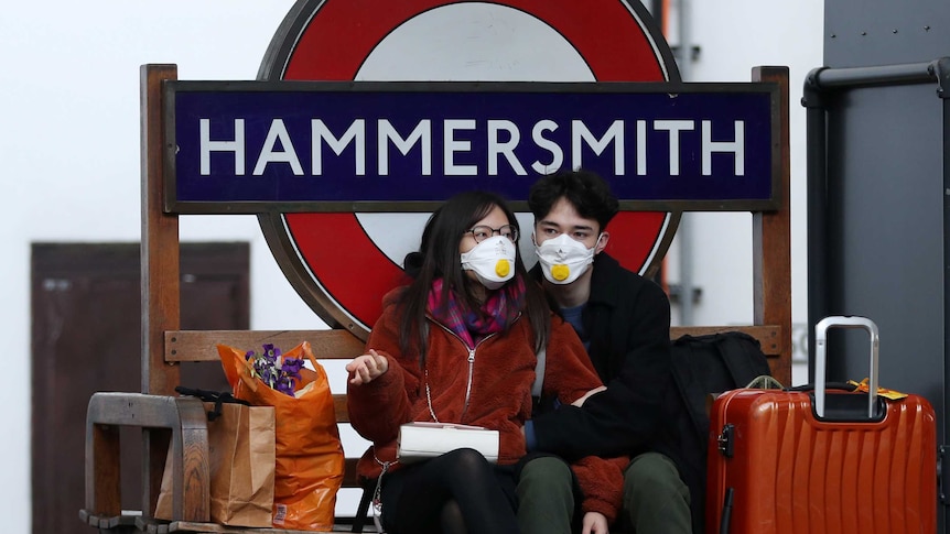 A couple in face masks leans against each other on a bench outside Hammersmith tube station