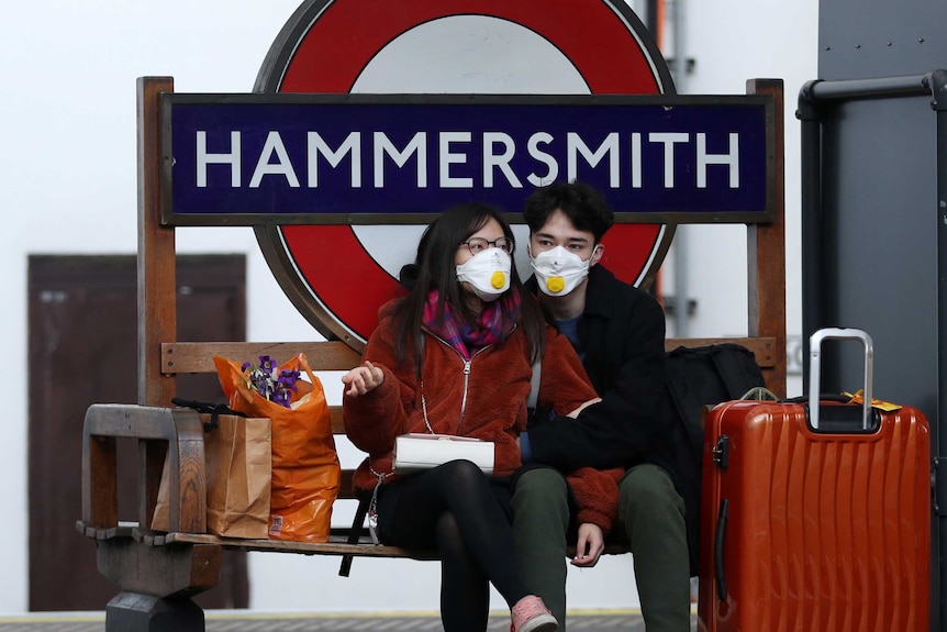 A couple in face masks leans against each other on a bench outside Hammersmith tube station