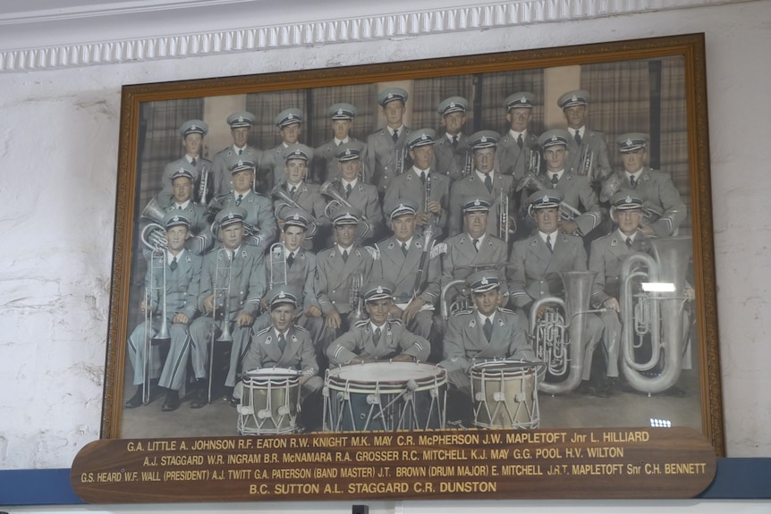 A painted photo of a band standing in a group in a wooden frame on a wall. There are three drums in front of the band 