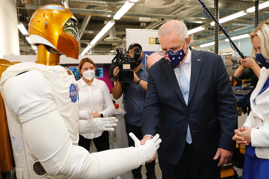Scott Morrison, wearing an Australian flag face mask, shakes hand with a robot.