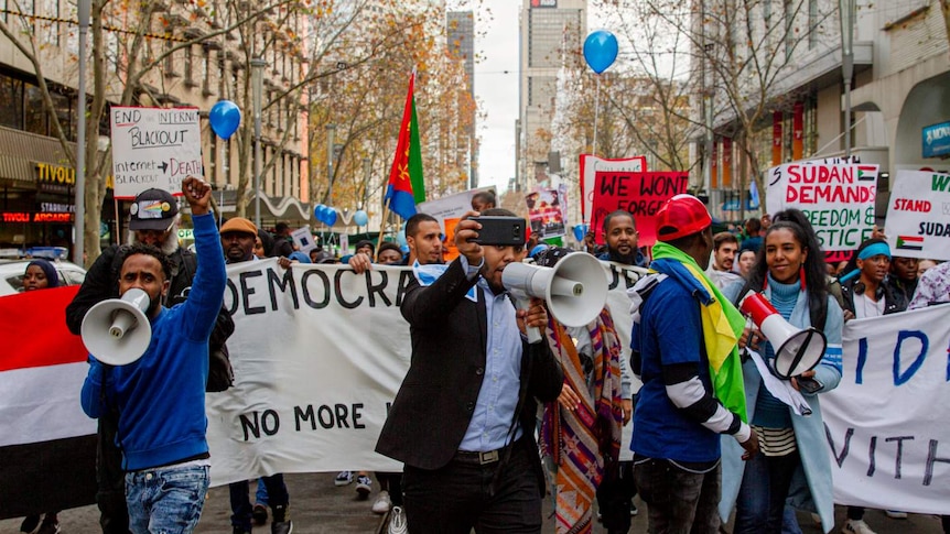 Looking down tramlines, you see a large crowd of Sudanese-Australian protestors with banners, megaphones, balloons and phones.