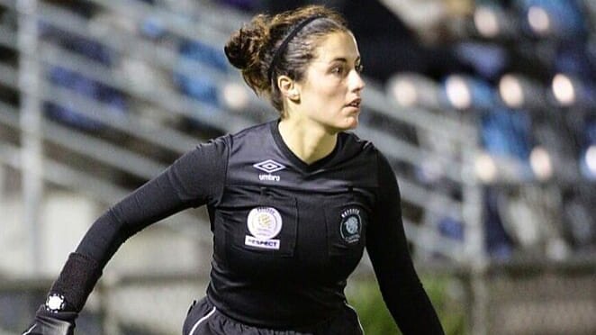 A woman, dressed in all black, running on a soccer field with a referee flag.
