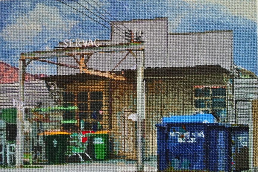 Needlepoint of rusted old warehouse with bins in front of it, and blue, cloudy sky above it.