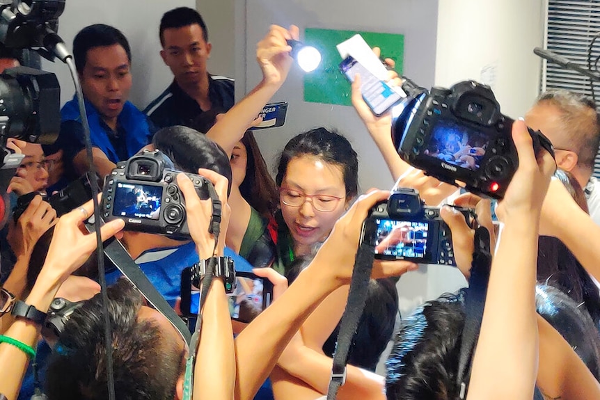 A woman with glasses is seen behind a throng of cameras she she holds up a flashlight to read a written statement.