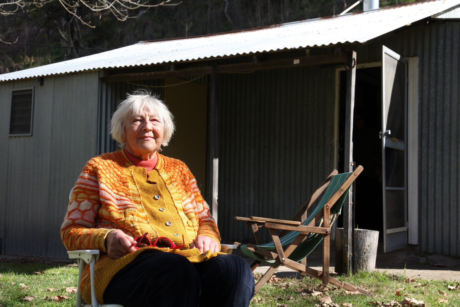 An elderly woman wearing a bright orange sweater sitting in a lounge chair in front of a corrugated iron shack.