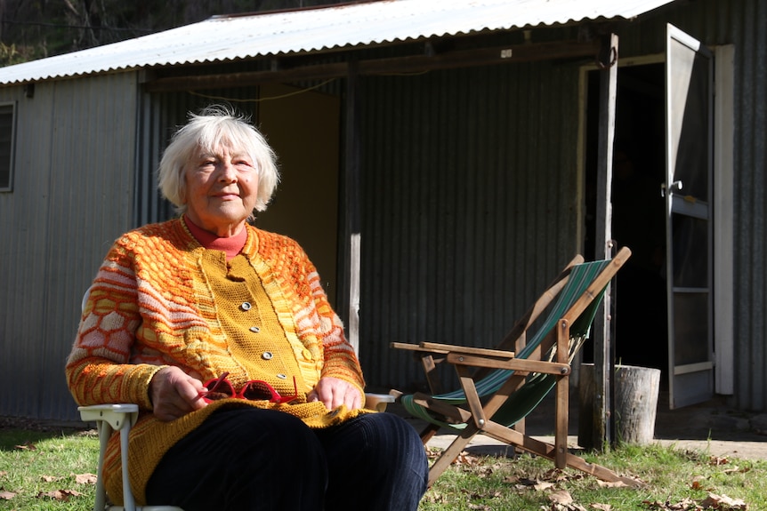 An elderly woman wearing a bright orange sweater sitting in a lounge chair in front of a corrugated iron shack.