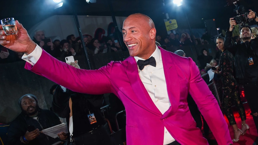 Dwayne the Rock Johnson wears a hot pink suit jacket and waves at a crowd at a film premier