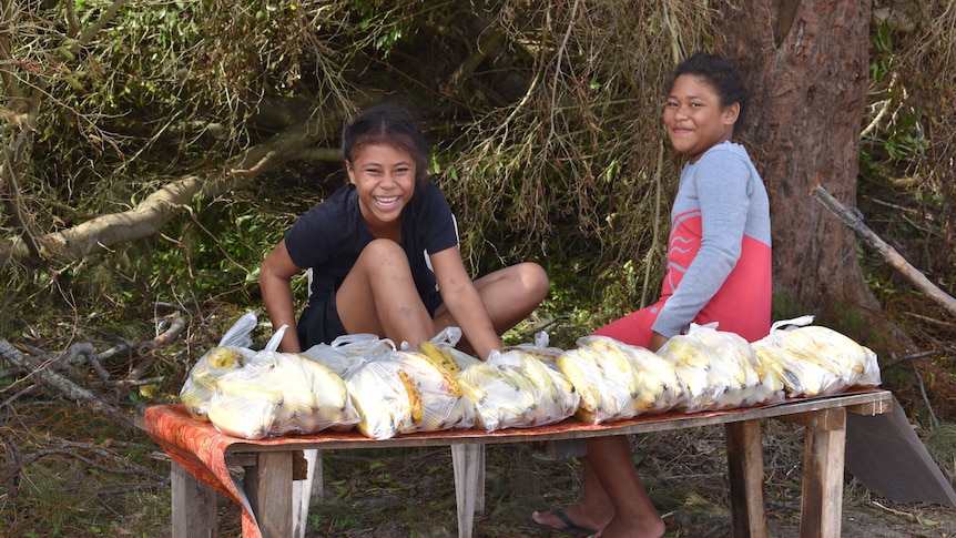 Two girls sit on a bench with bags of bananas.