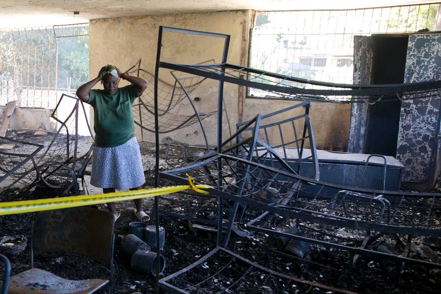 A woman stands with her hands on her head as she stands on blacked rubble and looks at burned bed frames.
