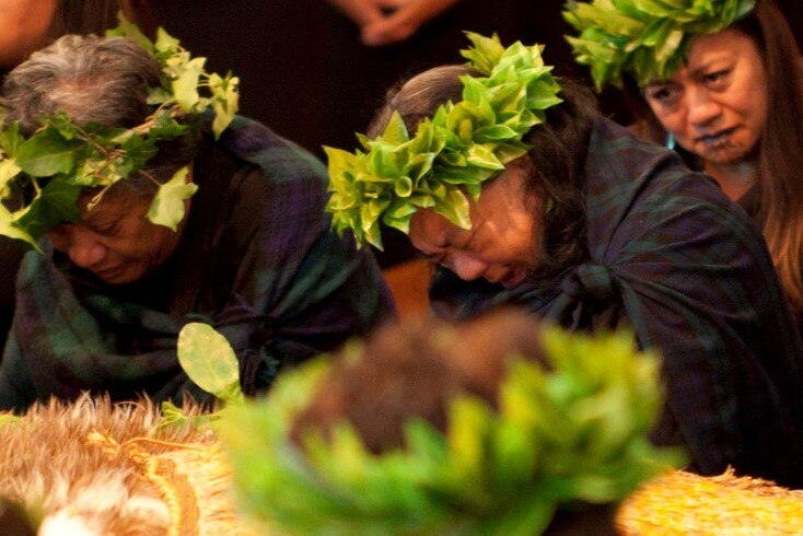 A group of women wearing black and leafy head-wreaths weep as they pay respects to the remains of a repatriated ancestor.