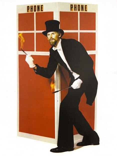 A man wearing a black tuxedo and top hat holds sticks burning with fire, in front of a phone box. 