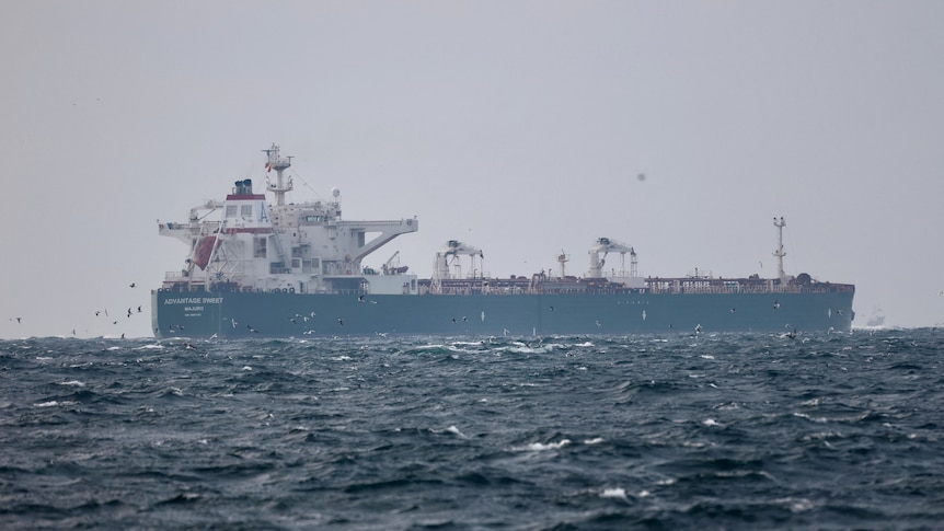 A blue and white oil tanker sails across a choppy sea with lots of birds flying around it.