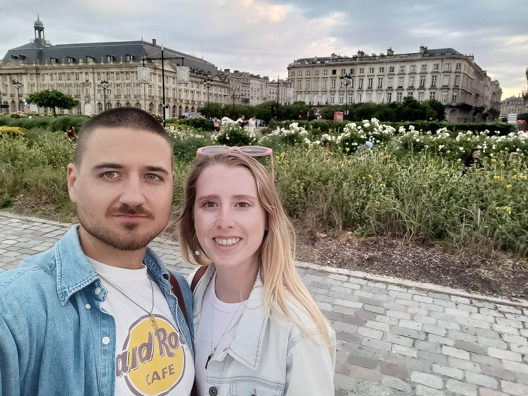 A man and woman stand together with a grand historical building in the background. She has long blonde hair.