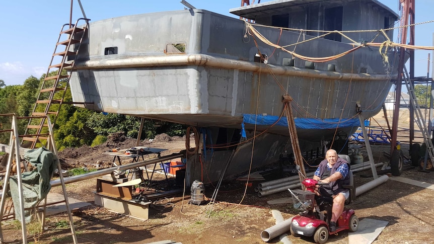 A man riding a red mobility scooter is dwarfed by a large boat under construction on land.