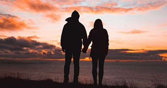 The silhouette of the backs of two people standing near ocean watching the orange clouds of a sun setting or rising.