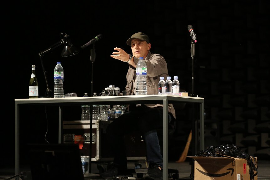 Simon McBurney, a middle-aged white man wearing a grey shirt and black cap, sits at a table giving notes in a theatre.