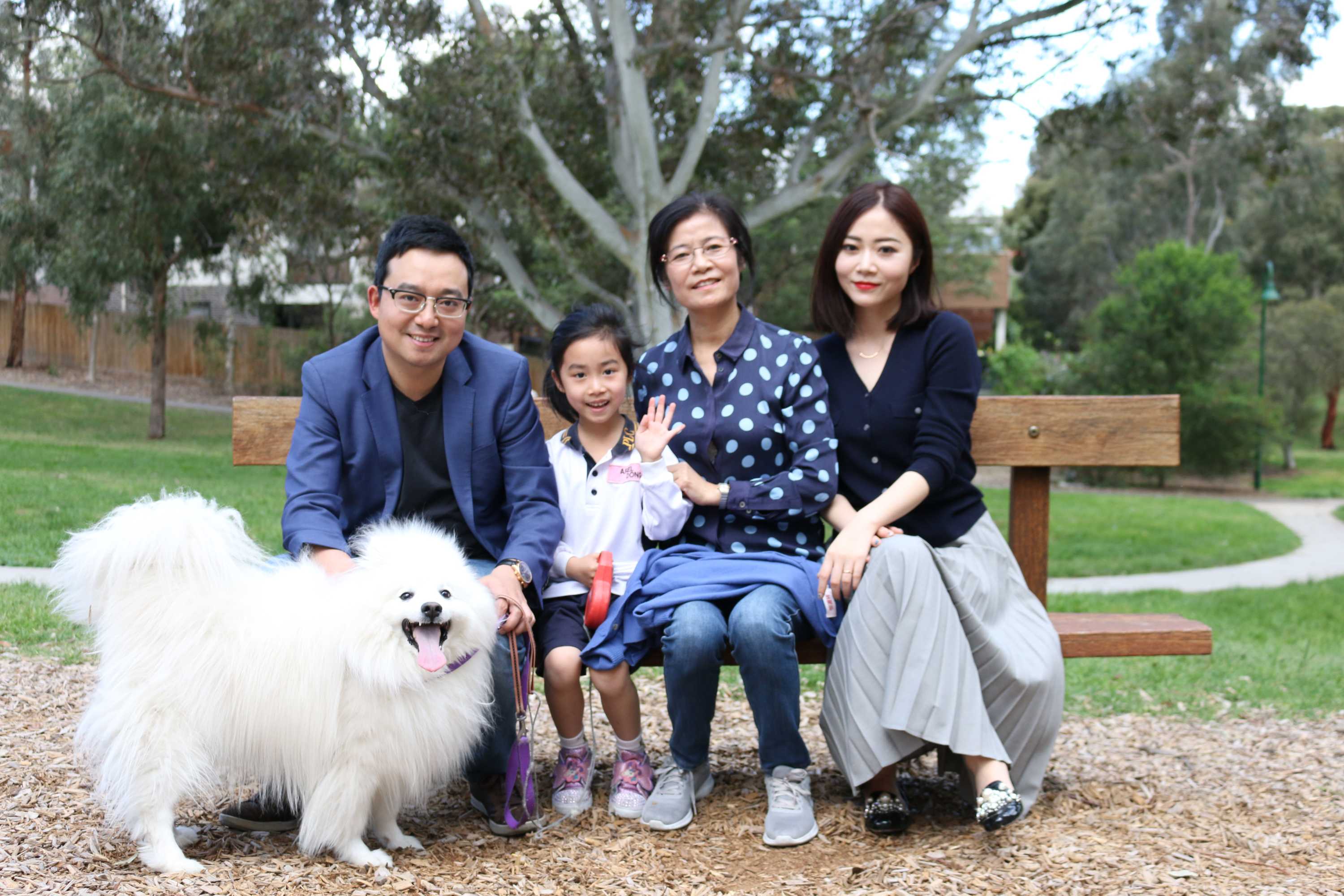 Sean Dong with his daughter, mother-in-law and wife sitting on a park bench smiling for a photo, with their pet dog in front.