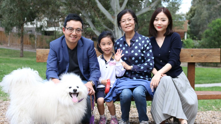 Sean Dong with his daughter, mother-in-law and wife sitting on a park bench smiling for a photo, with their pet dog in front.