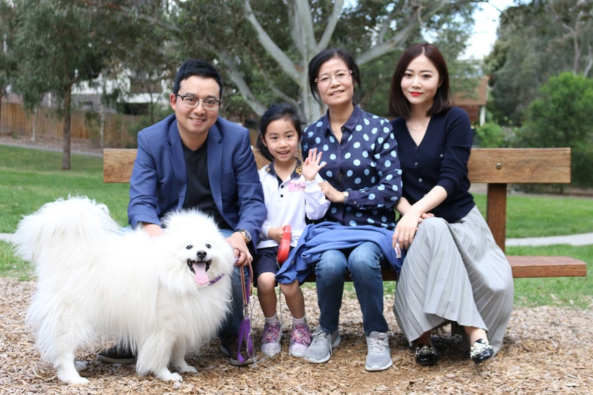 Sean Dong with his daughter, mother-in-law and wife sitting on a park bench smiling for a photo, with their pet dog in front.