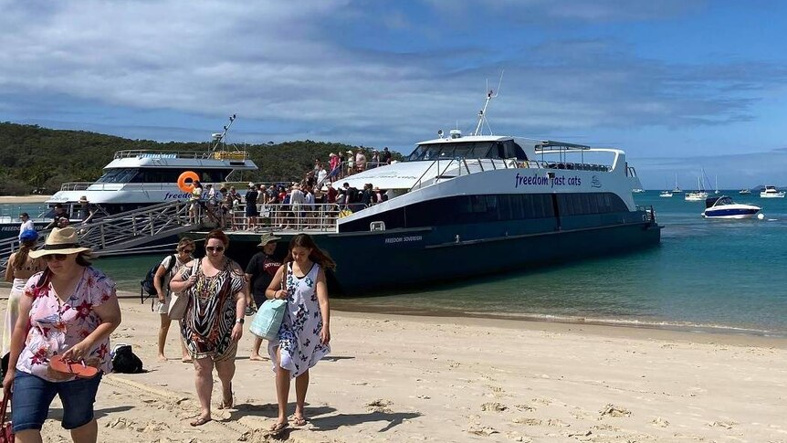 A crowd of people walk down a ramp on a ferry to a beach.