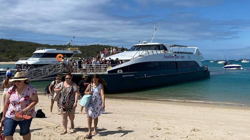 A crowd of people walk down a ramp on a ferry to a beach.