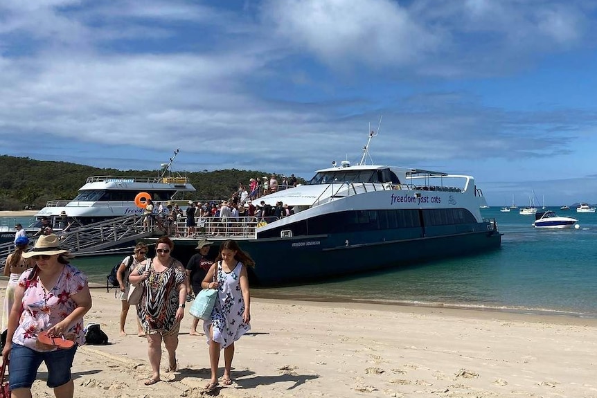 A crowd of people walk down a ramp on a ferry to a beach.