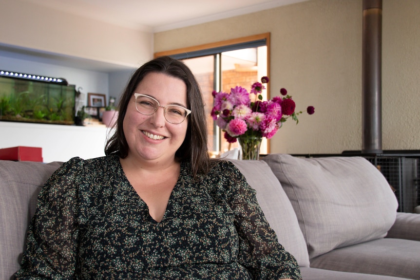 A smiling woman with glasses is sitting on a couch in a living room