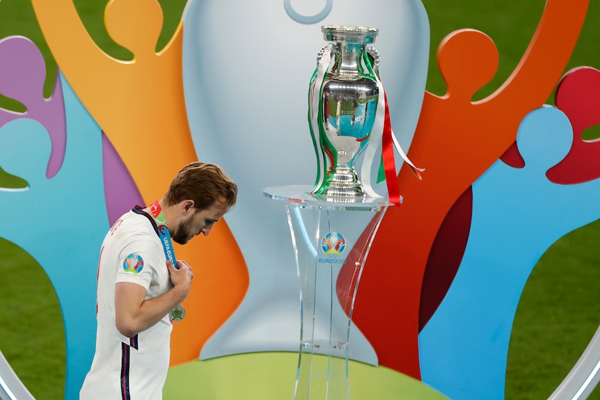 Harry Kane walks past a large silver trophy in front of the colourful backdrop