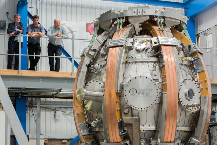 Three men elevated walkway looking at a large round mechanical device that appears to be made of metal and copper.