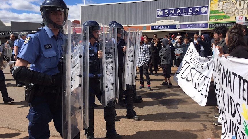 Police in riot gear with shields and helmets line up opposite protesters carrying banners on Hannan Street in Kalgoorlie.