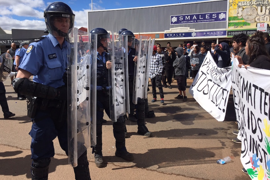 Police in riot gear with shields and helmets line up opposite protesters carrying banners on Hannan Street in Kalgoorlie.