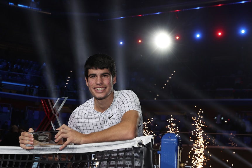 A young Spanish tennis player smiles at the net as he holds a tournament trophy.