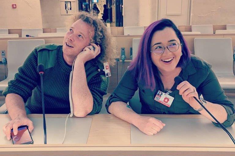 Two donor-conceived people sit at a bench in a convention room in front of microphones smiling and posing for the camera.