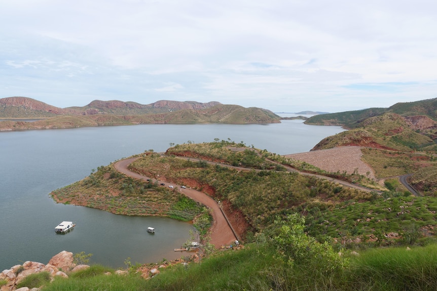 Image of a freshwater lake, surrounded by hills. There are boats in the foreground and a road winding around the cliffs.